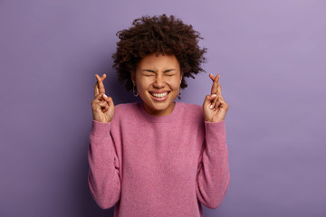 Pleased emotive cheerful woman prays God for good luck, crosses fingers, smiles broadly, shows white teeth, wears rosy jumper, gestures against purple background, waits something positive happen