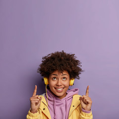 Glad positive woman has curly hairstyle, points both index finger on ceiling, shows copy space above, has good mood as listens amusing songs, wears stereo headphones on ear. Look upwards, please