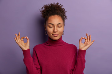 Portrait of good looking woman meditates, keeps both hands in okay gesture, keeps eyes closed, practices yoga to relax after work, wears burgundy jumper, isolated on purple wall. Zen posture