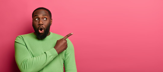 Shocked emotive bearded man with black skin, wears bright green sweater, points at empty space,...