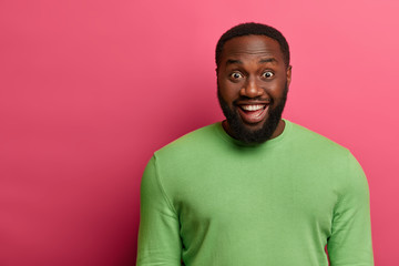 Fototapeta premium Handsome bearded man has thick bristle, looks joyfully at camera, hears something very pleasant while talks with colleague, wears neat green jumper, isolated on pink wall with copy space left