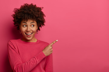 Positive delighted woman with Afro haircut points index finger at upper right corner, recommends...