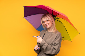Young blonde woman holding an umbrella over isolated yellow wall pointing finger to the side