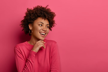 Isolated shot of curly ethnic woman holds chin, turns head aside, laughs positively, has healthy dark skin, enjoys spending nice free time in merry company, wears bright clothes. Monochrome. Emotions