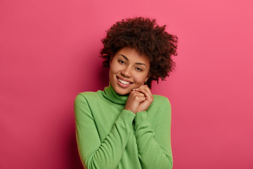 Fototapeta na wymiar Portrait of good looking curly woman tilts head, feels pleased to spend weekend with her family, keeps hands presed together, wears green jumper, shows white teeth, expresses positive emotions