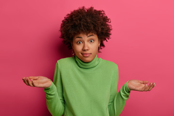 Indifferent confused black woman spreads palms confusingly, has doubts, wears green turtleneck, has...