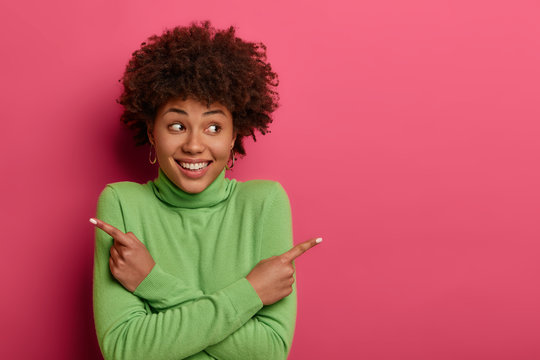 Cheerful curly haired woman crosses arms over chest, points left and right, gives two variants, says both are good, advertises products, wears green turtleneck, stands against crimson background