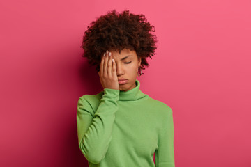 Fototapeta na wymiar Photo of tired Afro American woman covers half of face with palm, keeps eyes closed, wants to sleep, needs rest, dressed in green sweater, isolated on pink background. Negative feelings concept