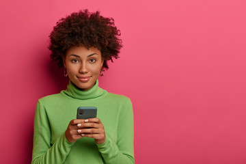 Good looking woman with Afro hair types message on smartphone, browses network, looks confidently at camera, has online communication via special application, wears green poloneck. Mobile services