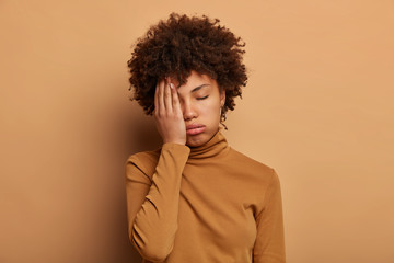 Fototapeta na wymiar Photo of tired curly woman covers face with palm, feels overworked and fatigue, wants to sleep, tilts head, wears casual turtleneck, isolated over brown background. Tiredness, dissatisfaction concept