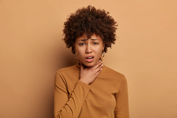 Fototapeta na wymiar Displeased dark skinned woman has sore throat, feels pain after cathcing cold, being sick and unwell, frowns face, wears brown jumper, stands indoor. Painful inflammation. Discomfort caused of illness
