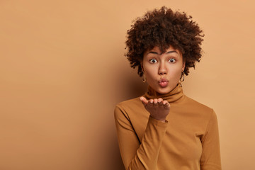 Fototapeta na wymiar Passionate good looking woman with Afro hairstyle blows airkiss, holds palm near folded lips, makes flity message to boyfriend, stands indoor against brown background. Romantic feeling, tenderness