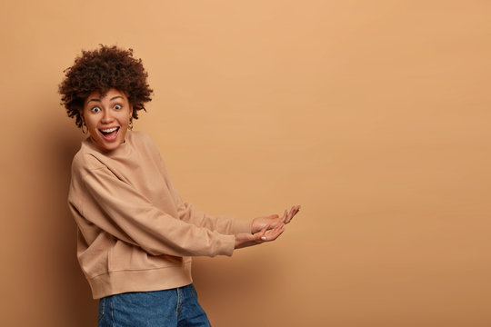 Happy African American woman stands sideways, pretends holding something from below, stands with palms raised, carries invisible object, dressed in casual wear, isolated on beige background.