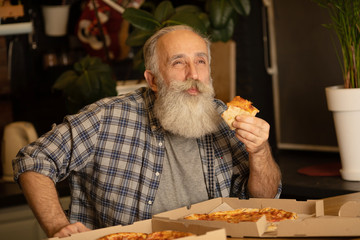 Smiling older man eating pizza slice sitting at living room. Bearded senior man eating italian food.