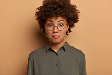 Fototapeta na wymiar Headshot of curly woman with wondered expression, expresses surprisement, hears intiguing news, wears round spectacles, grey shirt, isolated over beige background. Facial expressions concept