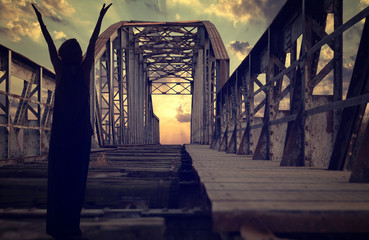 Silhouette of person with raised hands praying on old abandoned railway metallic bridge with the sunset in the background