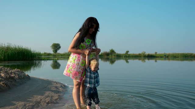  Young Beautiful Mother And Her Child Play And Walk Near River. Summer Lake Morning Sunny Day Blue Sky. Slow Motion 30p 0.5 60p