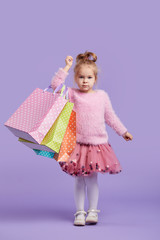 Portrait of a happy little child girl standing on a purple background holding shopping bags, package. Looking at the camera.
