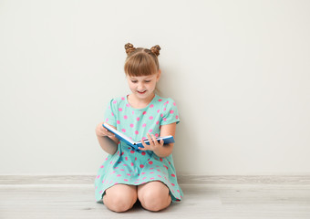 Cute little girl with book sitting near white wall