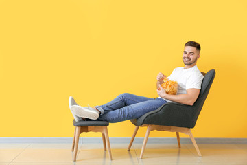 Handsome young man with tasty potato chips sitting in armchair near color wall