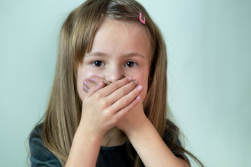 Close-up portrait of little child girl with long hair covering her mouth with hands.