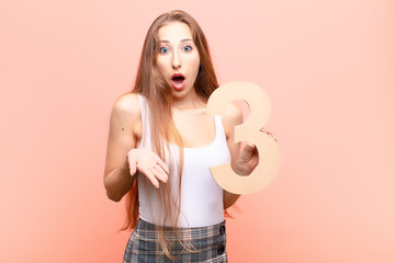 yound blonde woman surprised, shocked, amazed, holding a number 3.