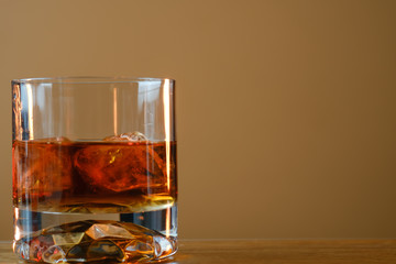 Glass of whiskey with ice cubes served on wooden board. Dark background, glossy golden color hard liquor on countertop with highlight. Night life bar relaxation concept