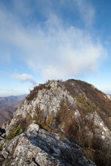 Rocky mountain peak after a narrow, rocky ridge, dry, late autumn vegetation and cloudy blue sky