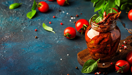 Italian Sun Dried tomatoes in olive oil with green basil and spices in glass jar on blue kitchen table, copy space