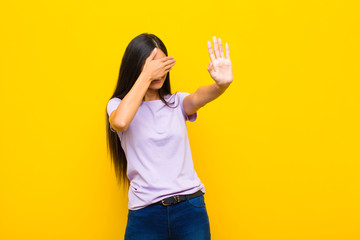 young pretty latin woman covering face with hand and putting other hand up front to stop camera,...