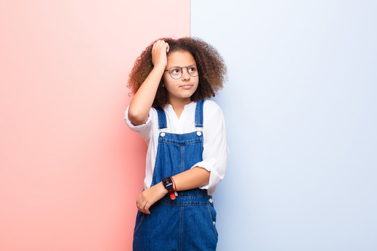 african american little girl feeling puzzled and confused, scratching head and looking to the side against flat wall