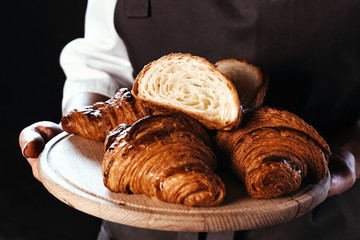 Many fresh croissants on wooden board in hands of baker. Dark background, cafe.