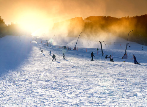 Ski resort, snowmaking on artificial slopes at sunset. Skiers and sheer snowboarders on the slope.