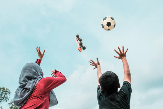 Rear View Of Siblings With Arms Raised Against Skateboard And Soccer Ball In Mid-Air