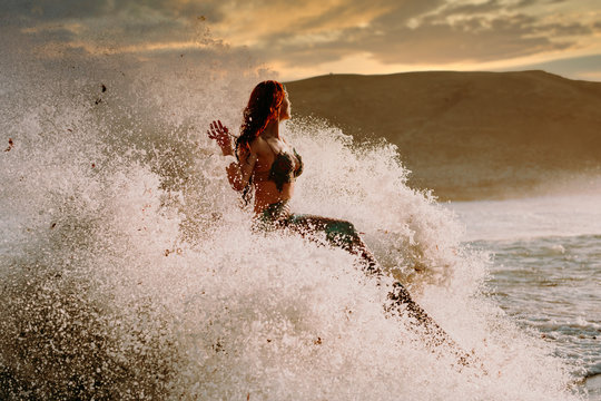 Caucasian redhead woman with mermaid tail perches on rock with pounding waves around her