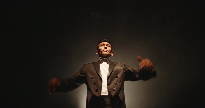 Asian male orchesra conductor wearing tux is directing symphony orchestra with movement of his hands and stick, studio shot on black background 4k footage