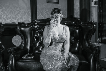 a girl in a gold dress with a diadem on her head, drinks from a glass, sits on a leather armchair, next to a gun and beads