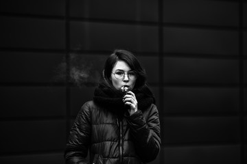 Vape teenager. Young pretty white caucasian girl in a jacket and glasses smoking an electronic...