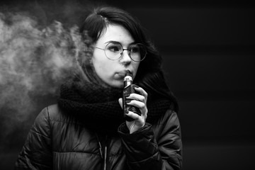 Vape teenager. Young pretty white caucasian girl in a jacket and glasses smoking an electronic...