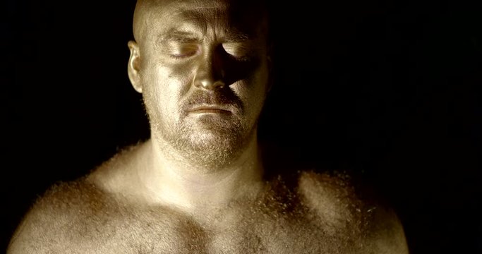 portrait of a bald unshaven man with a hairy body and shining Golden skin on a black background. he stands with his eyes closed, then opens them, screams and shows his teeth