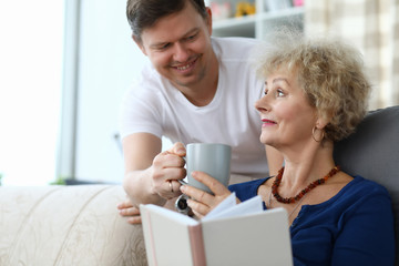 Portrait of smiling man giving cup of tea to mom. Woman sitting on sofa and reading book. Family spending time together. Warm relationship concept