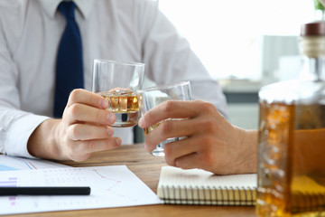 Close-up of male hands holding glasses with whiskey. Partners celebrating signing important agreement in office. Colleagues relaxing after busy day. Business negotiations concept