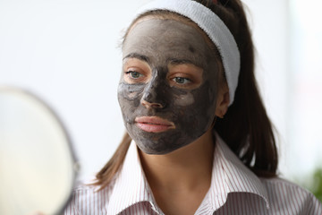 Close-up of charming young woman looking in mirror and applying wetting facial clay mask. Beautiful female relaxing during skin therapy. Wellness and spa concept