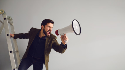 A bearded businessman on the stairs with a megaphone in hand shouts on a gray background.