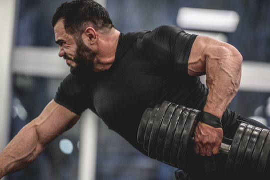 strong young bearded caucasian man in black sportswear lifting heavy weight dumbbell with one muscle with vein hand training back and arms in sport gym training routine