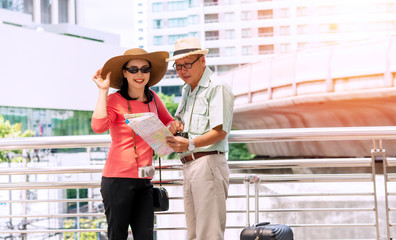 Traveler couple elderly wear hat with luggage, camera and binoculars holding map in hands while walking together in street city. They are looking map on vacation pointing for some destinations.