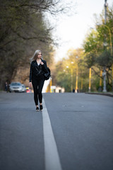 A young beautiful blonde girl in a black leather jacket is walking along an empty city road, the lower angle of the shooting