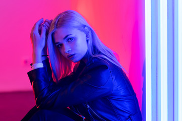 Young beautiful blonde girl in black leather jacket in red and blue light sitting on the floor