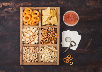 Glass of craft lager beer and opener with box of snacks on wooden background. Pretzel,salty potato sticks, peanuts, onion rings with nachos in vintage box with openers and beer mats.