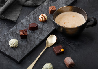 Luxury Chocolate candies selection with cup of cappuccino coffee and dessert spoon on black marble board and dark table background.